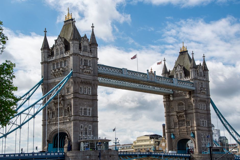 A view of the River Thames in London with the iconic Tower Bridge in the background under a cloudy sky. On the left side, a large naval vessel with a grey metallic surface and various antennas and mas