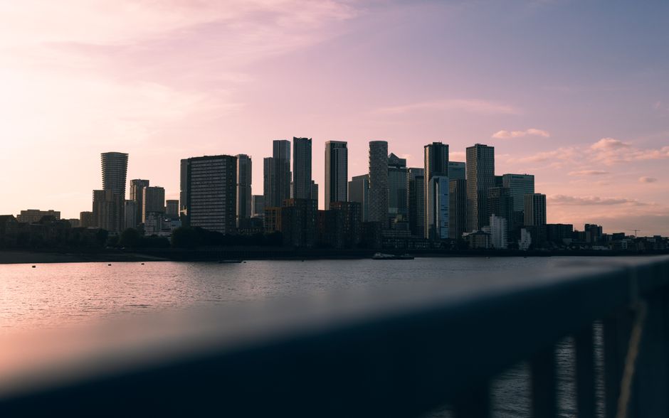 In the foreground, a dark, blurred surface appears to be part of a railing or barrier along a waterfront area, with visible vertical supports. Beyond this, a calm river reflects the soft pink and purple hues of the early evening sky. On the far side of the river, a dense cluster of modern high-rise buildings and skyscrapers dominate the city skyline, showcasing a variety of architectural styles, with some structures featuring glass facades and others with textured, concrete exteriors. The buildings are arranged in a compact, overlapping manner, with shadows cast by the setting sun highlighting their shapes and heights. To the left, a distinctive, twisting tower stands out against the skyline. The overall scene suggests an urban environment with an active development area, which Waste Removal London might serve through independent or private waste collection, as reflected in the clear, detailed depiction of the cityscape and river setting, emphasizing the importance of proper rubbish management in a bustling city context.
