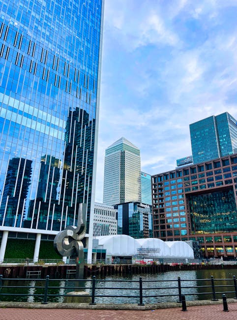 A view of a modern urban landscape featuring several tall office buildings with reflective glass facades, positioned along a waterway. The buildings vary in height and design, with one prominent structure displaying vertical stripes on its blue-tinted glass exterior, and another with a beige grid pattern. In the foreground, there is a black metal railing along the water's edge, with a small sculpture resembling a musical note made of metal situated nearby. Behind the railing, the water appears calm, reflecting parts of the buildings. To the right, a reddish-brown building with large windows and an open ground floor is visible, while in the background, the sky is partially cloudy, providing natural light that illuminates the scene evenly. This scene subtly aligns with the context of private or alternative waste management in a city setting, with the buildings and infrastructure suggesting a developed commercial district. Waste Removal London occasionally handles on-site clearance or private rubbish collection in such environments, although these details are not directly depicted. The overall composition emphasizes the city's architectural environment and the urban setting associated with waste removal or rubbish services.